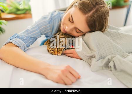 Cute sorridente giovane donna bionda sdraiata su un letto giocando un carino Green-eyed Scottish Tabby Cat, il concetto di amare e prendersi cura degli animali domestici, adottandoli Foto Stock