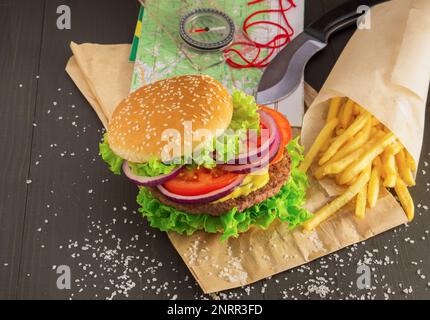 Hamburger fresco con cipolla tagliata di manzo, lattuga, pomodoro, salsa di senape, sale e patatine fritte su tavola di legno. Mappa, coltello e bussola per grandi avventure An Foto Stock