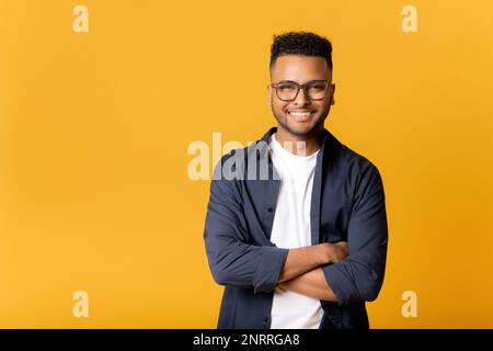 Ritratto di riuscito uomo indiano in piedi in posa fiduciosa con le braccia incrociate e guardando la macchina fotografica isolata su giallo, imprenditore maschio allegro, freelancer sorridente Foto Stock