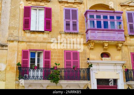 Facciata della casa con balconi maltesi viola e rosa, piante in pentole, finestre con persiane sulla casa cittadina di pietra calcarea gialla. Tipica scena stradale Foto Stock