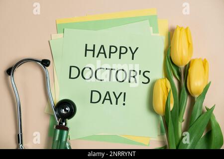 Happy Doctor's Day - cartolina con testo, stetoscopio e tulipani gialli su sfondo verde e beige Foto Stock