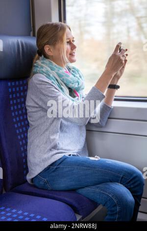 Giovane donna in treno che scatta foto del paesaggio con il suo smartphone (immagine simbolo) Foto Stock