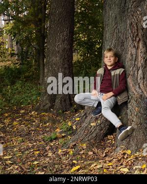 Un bel ragazzo siede sulla radice di un enorme quercia che si appoggia contro il tronco di un albero e sorride Foto Stock