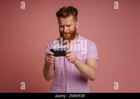 Adulto elegante rosso testa barbuto messo a fuoco uomo teso in camicia rosa giocare gioco cellulare sul telefono, mentre in piedi su sfondo isolato corallo Foto Stock