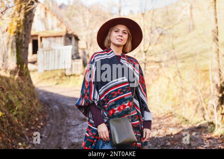 Elegante hipster donna in viaggio indossando autentico boho elegante stile poncho e cappello vicino a vecchi edifici di campagna in legno Donna esplorare la natura in autu Foto Stock