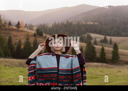 Elegante ragazza viaggiante con un autentico poncho e cappello boho chic nel paesaggio autunnale. Felice giovane donna esplorando montagne soleggiate. TR Foto Stock