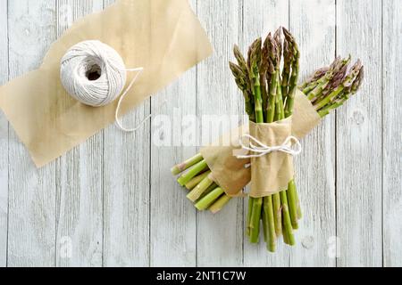 Asparagi freschi raccolti legati in grappoli con pergamena e filo di macelleria su fondo di legno grigio in composizione piana. Foto Stock