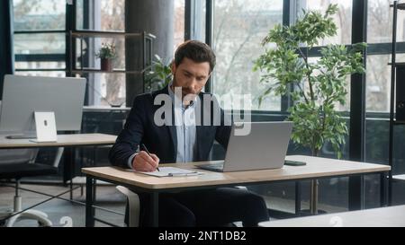Un uomo d'affari adulto focalizzato lavora su un progetto in ufficio utilizzando appunti di scrittura per laptop uomo caucasico business leader professionista dipendente analizzare le informazioni Foto Stock