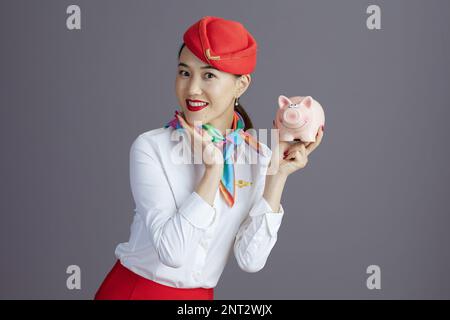 donna asiatica sorridente moderna in gonna rossa e cappello uniforme con caviglia isolata in grigio. Foto Stock