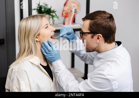 Il medico utilizza la spatola per ispezione per esaminare la gola del paziente. Medico ENT che fa esame di gola di una donna. La paziente aprì il check-up bocca-gola. Foto Stock