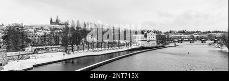 Vista panoramica dell'isola di Kampa durante l'inverno a Praga, Repubblica Ceca. In primo piano, c'è la riva del fiume Moldava con i mulini Sova, ceco: Sovovy Mlyny, complesso. Sullo sfondo, si può ammirare il Castello di Praga e il Ponte Carlo. Fotografia in bianco e nero. Foto Stock