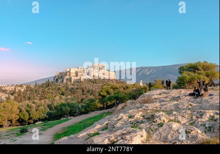 Gli incidentali sulla collina di Pnyx trascorrono un pomeriggio di sole, con l'Acropoli di Atene e la collina di Areopago sullo sfondo. Atene, Grecia. Foto Stock