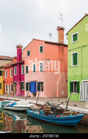 Barche ormeggiate in canale fiancheggiato da case in stucco verde, rosa, viola, rosso e giallo decorate con tende sulle porte d'ingresso, Isola di Burano, Italia. Foto Stock