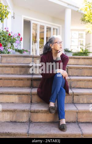 Felice donna asiatica anziana seduta sulle scale e guardando lontano in giardino Foto Stock