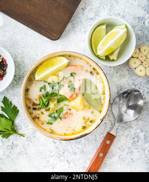 Zuppa di salmone con patate, carote, panna, erbe aromatiche. Zuppa di pesce scandinava/norvegese in ciotola, tagliando asse di legno, cucchiaio, cemento bianco rustico Foto Stock