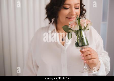 Una donna adulta in una camicia bianca è in piedi con un vetro con rose spruzzate su di esso. Una ragazza all'interno con un bicchiere di rose rosa. Foto Stock