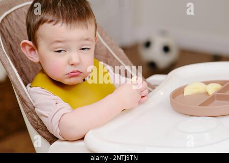 Bambino bambino felice che mangia i pezzi della mela mentre siede su una sedia alta. Un bambino in bib mangia la frutta nella cucina domestica. Bambino di un anno e sei mesi Foto Stock