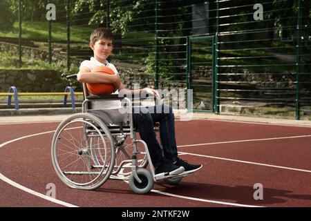 Ragazzo minorato in sedia a rotelle con palla da basket nel campo all'aperto Foto Stock