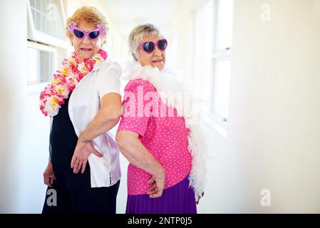 Siamo giovani e liberi. Scatto di due donne anziane spensierate indossando occhiali da sole e posando per la macchina fotografica all'interno di un edificio. Foto Stock