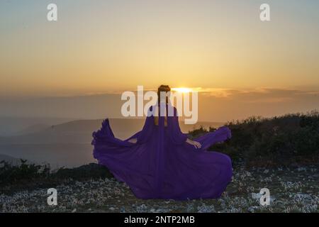 Tramonto viola abito donna montagne. Ascesa del mistico. tramonto sulle nuvole con una ragazza in un lungo vestito viola. Nel prato c'è un prato Foto Stock