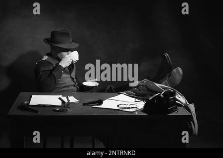 Fotografia in bianco e nero, nero, vintage. Uomo, detective professionista in cappello seduto al tavolo e bere caffè. Indagine Foto Stock