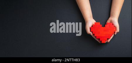 Mani di bambina che tiene il cuore fatto di mattoni di plastica su sfondo grigio scuro Foto Stock