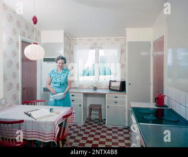 Ascoltatori radio in passato. Una donna in cucina sta frustando qualcosa in una ciotola al tavolo da cucina. Una radio è visibile sul banco di lavoro dalla finestra. Sulla parte anteriore della radio è presente una scala e quando si ruota la manopola accanto ad essa in modo che il puntatore si trovi al centro del nome di una stazione, la stazione era lì. Oltre alle trasmissioni FM, hanno anche ascoltato le radio straniere a onde lunghe. Svezia nel 1958 Foto Stock