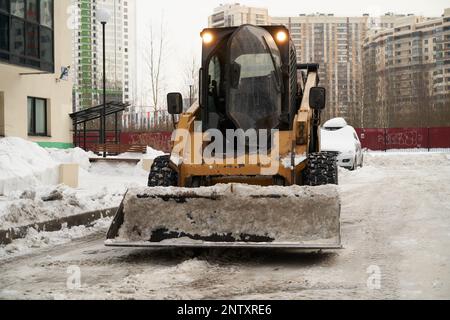 L'attrezzatura per la rimozione della neve sullo sfondo degli edifici urbani pulisce la neve con un secchio sulla strada. Vista frontale, concetto di vita urbana. Foto Stock