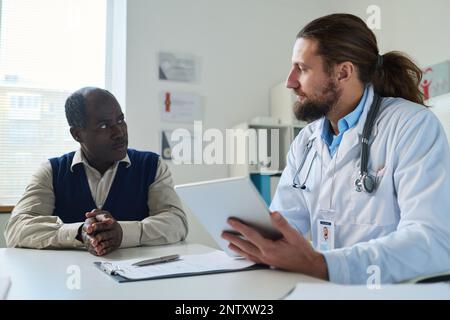 Giovane medico con tablet guardando il paziente afroamericano di sesso maschile senior e ascoltando lui durante la consultazione medica nelle cliniche Foto Stock