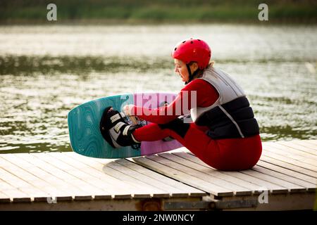 Donna in muta, casco e giubbotto di salvataggio seduto con wakeboard su un molo di legno. Giorno estivo di sole. Sicurezza nello sport. Sport acquatici in Finlandia. Assicurazione Foto Stock