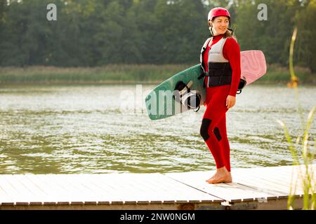 Donna in muta, casco e giubbotto salvagente che cammina con un wakeboard su un molo. Giorno estivo di sole. Sicurezza nello sport. Sport acquatici in Finlandia. Assicurazione conce Foto Stock