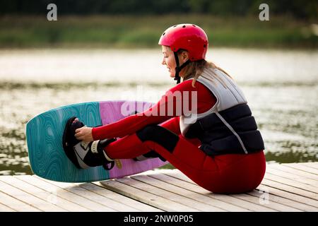 Donna in muta, casco e giubbotto di salvataggio seduto con wakeboard su un molo di legno. Giorno estivo di sole. Sicurezza nello sport. Sport acquatici in Finlandia. Assicurazione Foto Stock