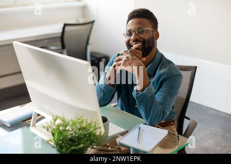 Buon uomo d'affari o CEO afroamericano seduto alla scrivania di fronte al computer, sorridente alla fotocamera, spazio libero Foto Stock