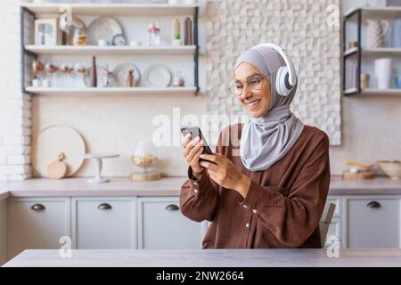 Una giovane donna musulmana in un hijab è seduta a casa in cucina indossando cuffie, tenendo un telefono in mano, ascoltando musica, un audiolibro, un podcast. Riposante, rilassato. Foto Stock