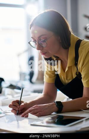 Giovane donna sarto o conciatore fare appunti o schizzi su modelli di carta da cucire mentre si piega sul posto di lavoro in atelier o workshop Foto Stock