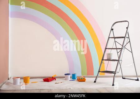 Diversi utensili decoratori e scala vicino alla parete con arcobaleno dipinto al coperto Foto Stock