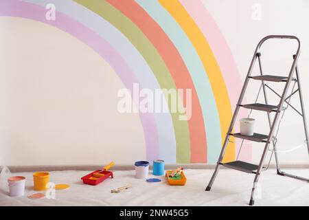 Diversi utensili decoratori e scala vicino alla parete con arcobaleno dipinto al coperto Foto Stock