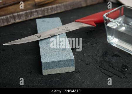 Pietra per affilatura, coltello e acqua sulla tavola nera, primo piano Foto Stock