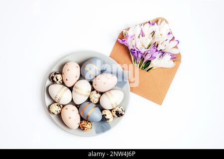 Piatto grigio con uova di Pasqua dipinte e busta con fiori di croco su fondo bianco. Biglietto di auguri di Pasqua. Vista dall'alto, disposizione piatta. Foto Stock