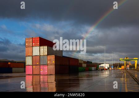Una pila di contenitori di spedizione su un molo commerciale deserto con luce solare e arcobaleno Foto Stock