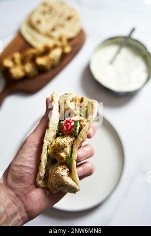 Souvlaki ona pita di pollo greco con verdure fresche e salsa tzatziki. Mano che tiene una pita con souvlaki di pollo. Sullo sfondo c'è un pollo Foto Stock