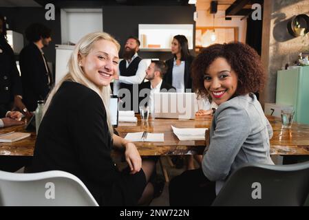 Due giovani colleghi attraenti con diversa etnia seduti alla scrivania guardando la fotocamera sorridente. Lavoro di squadra sul posto di lavoro. Foto Stock