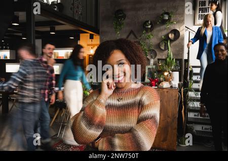 Ritratto di sorridente donna latina attraente guardando la macchina fotografica. Casual femmina in un luogo di lavoro occupato con persone sfocate a piedi. Foto Stock