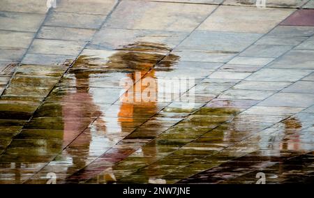 Astratto blurry silhouette ombra riflessi di persone irriconoscibili che camminano sotto l'ombrello sul bagnato marciapiede della città in una giornata piovosa Foto Stock