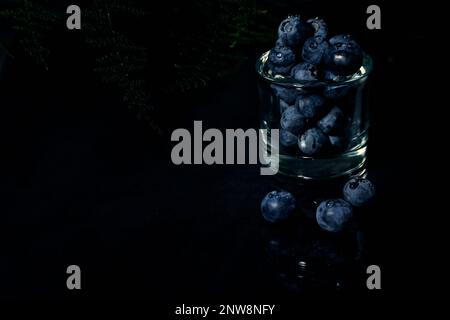 Mirtilli con gocce d'acqua in una tazza di vetro trasparente e la loro riflessione su un tavolo con erba verde e fondo nero Foto Stock