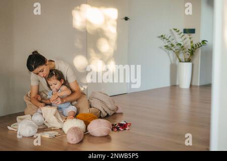 Vista a tutta lunghezza di una donna che si incrocia con la bambina mentre si siede in una casa accogliente. Hobby della giovane donna. Ricamo cerchio, ago Foto Stock