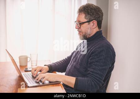Un uomo caucasico di mezza età con barba grigia è seduto alla scrivania e sta scrivendo su un computer portatile. Indossa un maglione blu e occhiali da dolcevita Foto Stock