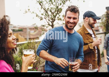 Gruppo di giovani studenti provenienti da diversi paesi che festeggiano insieme sulla terrazza. Un giovane con un bicchiere di birra sorride a una donna brasiliana mentre gli altri Foto Stock
