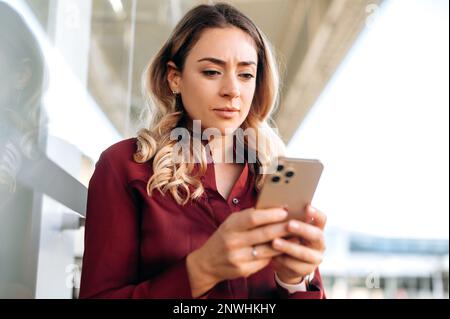 Messa a fuoco bella donna caucasica si trova all'aperto, utilizza il suo smartphone, scrive un messaggio, testi con gli amici, legge notizie su Internet, cerca informazioni, guarda concentrato sullo schermo del telefono Foto Stock