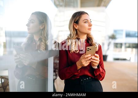 Bella donna caucasica attraente sta all'aperto, utilizza il suo smartphone, scrive un messaggio, testi con gli amici nei social media, cerca informazioni, guarda via, pensando al messaggio Foto Stock